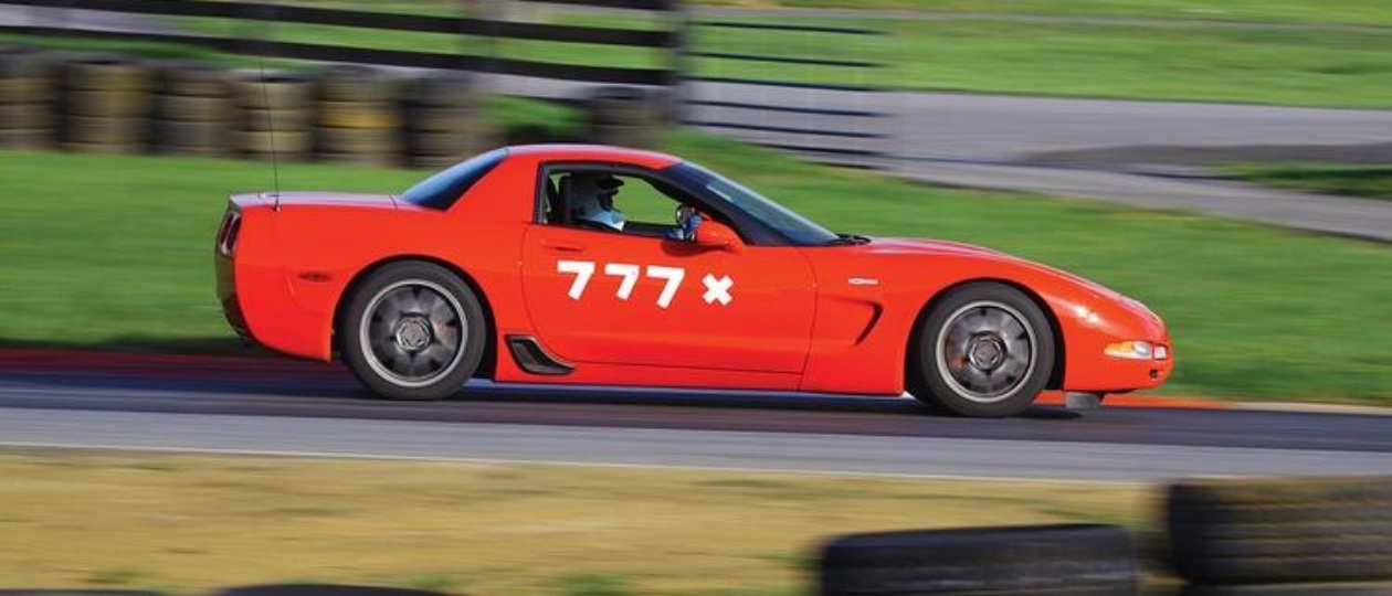 A red C5 Corvette Z06 parked on a race track with empty grandstands in the background
