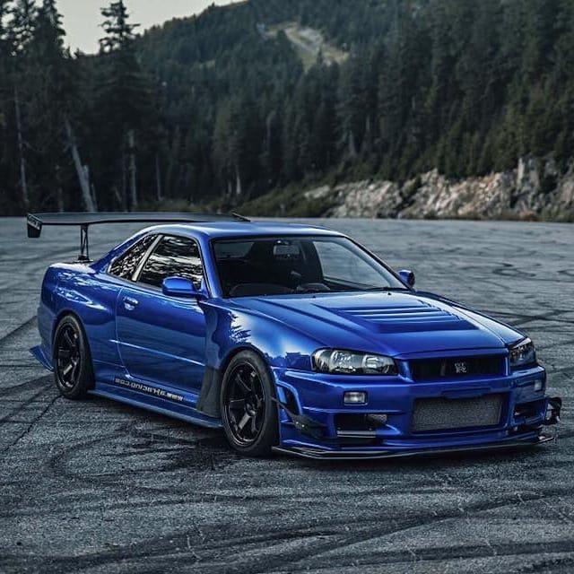 A Bayside Blue Nissan R34 Skyline GT-R with rear wing parked on mountain tarmac with pine forest behind